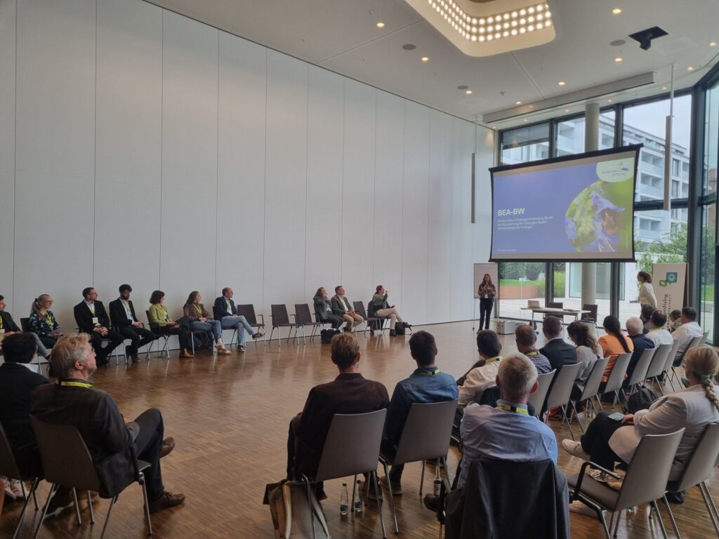 Saskia Wolf steht vorne neben ihrer Präsentation. Vor ihr sitzen Zuhörer*innen im Halbkreis. Der Raum ist hoch und lichtüberflutet.