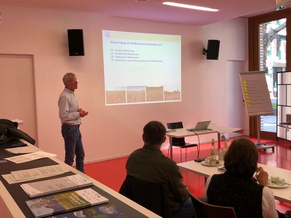 Ein Mann steht in einem Konferenzsaal vorne neben seiner Präsentation. Rechts im Bild sind zwei Zuhörer zu sehen.