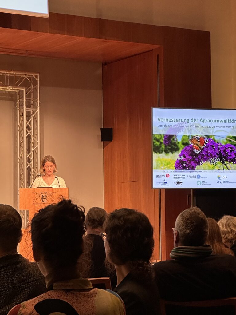 Eine Frau steht am Rednerpult. Rechts davon ist ihre Präsentation zu sehen. Im Vordergrund sind Hinterköpfe ihrers Auditoriums zu sehen.