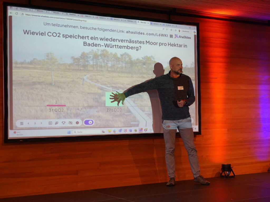 Volker Kromrey steht auf dem Podium und präsentiert seine Folien. Die Folie ist betitelt mit: Wieviel CO2 speichert ein wiedervernässtes Moor pro Hektar in Baden-Württemberg.