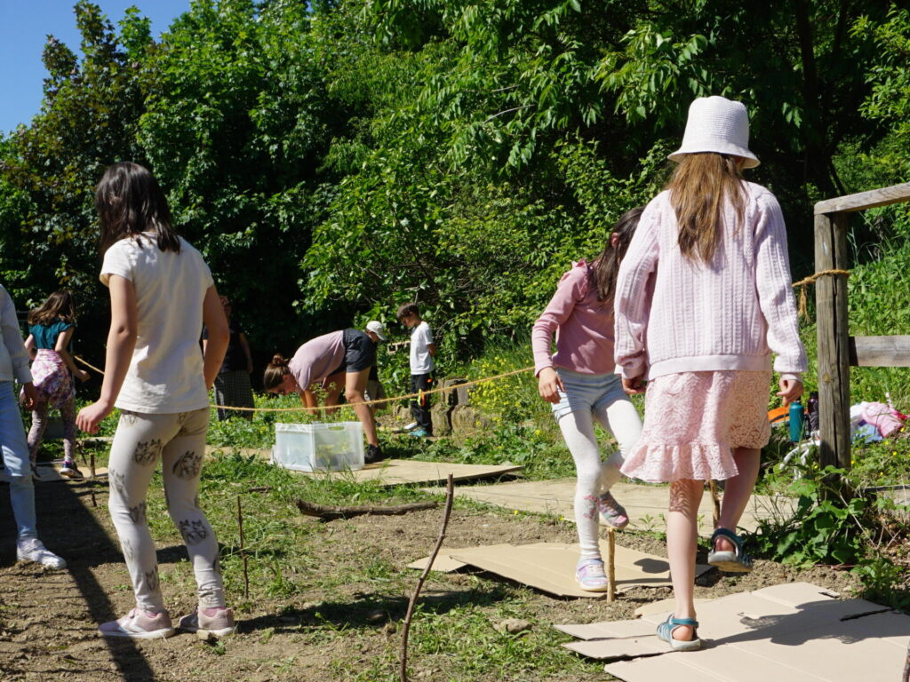 Kinder in Sommerkleidung stampfen im Freien bei Sonnenschein auf Kartons. Darunter liegen die frisch ausgebrachten Samen.