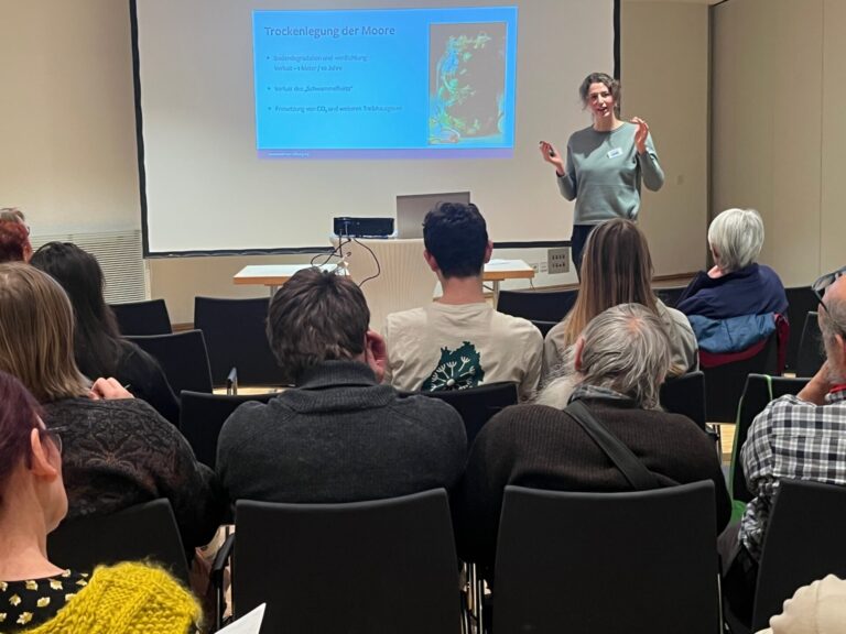 Luisa Gedon referiert vor einer Leinwand und einem zuhörenden Publikum, das in Kinobestuhlung vor ihr sitzt.