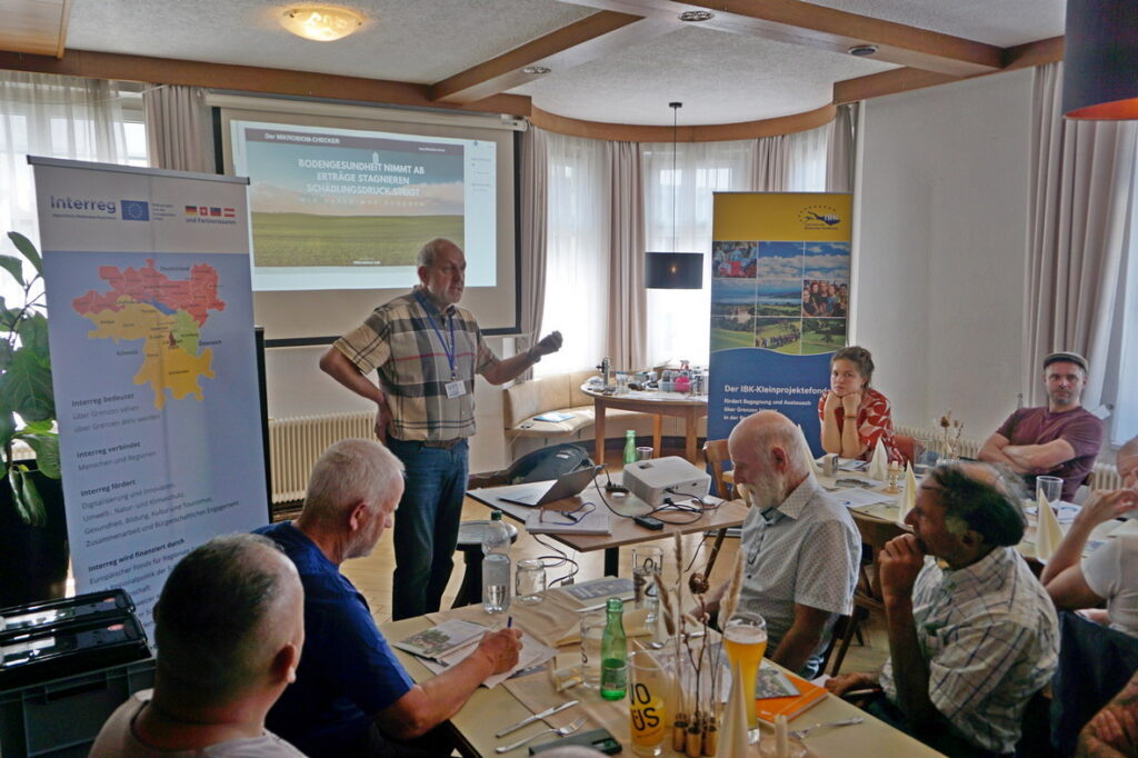 Peter Flaßhoff-Gockel steht vor seiner Präsentation über Bodengesundheit. Vor ihm sitzen Teilnehmer*innen an Tischen, teilweise mit Notizzettel und Stift. An den Seiten stehen Rollups von Interreg und IBK.