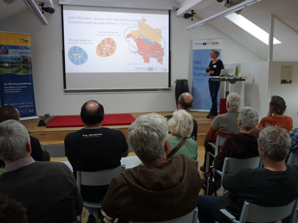 Blick von hinten in einen Vortragsraum. Zu sehen sind die Rücken der Zuhörenden, die auf die Referentin bzw. ihre Präsentation blicken.