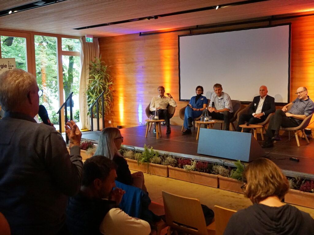 Zu sehen ist ein Podium, auf dem fünf Personen Platz genommen haben und gemeinsam mit dem Publikum über energierelevante Themen diskutieren. Im linken Vordergrund steht ein Mann mit Mikrofon, der einen Beitrag in das Plenum einbringt. Das Podium wird im Hintergrund von indirektem Licht aus Strahlern in warmen Orange- und kühlen Blautönen stimmungsvoll beleuchtet.