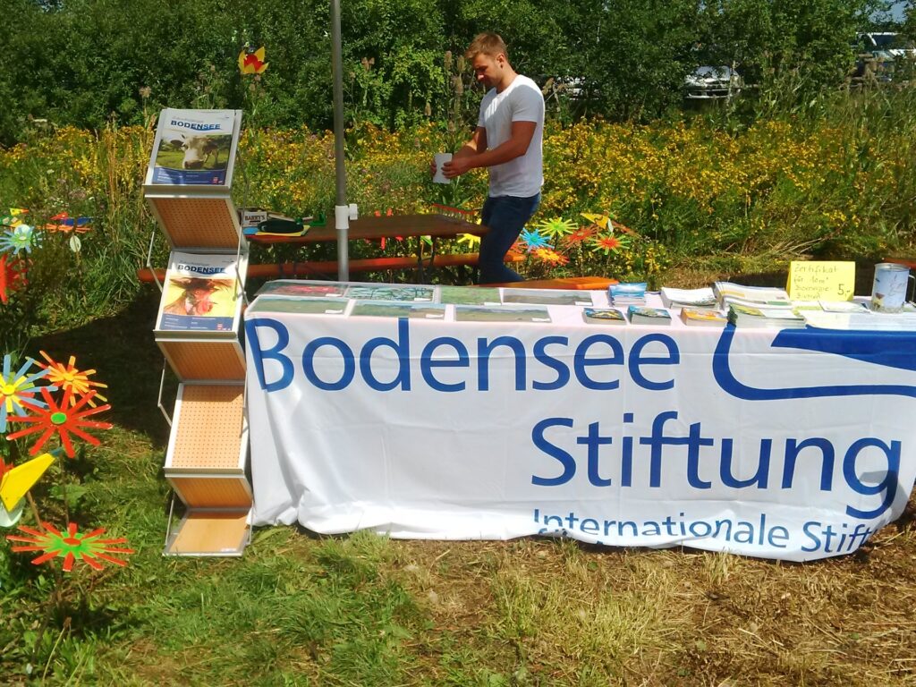 Info-Stand im Grünen: Ein Tisch ist mit einer Fahne und Informationsmaterial bedeckt. Auf der Fahne steht Bodensee-Stiftung. Hinter dem Tisch steht ein Mann. Er legt weiteres Material aus.
