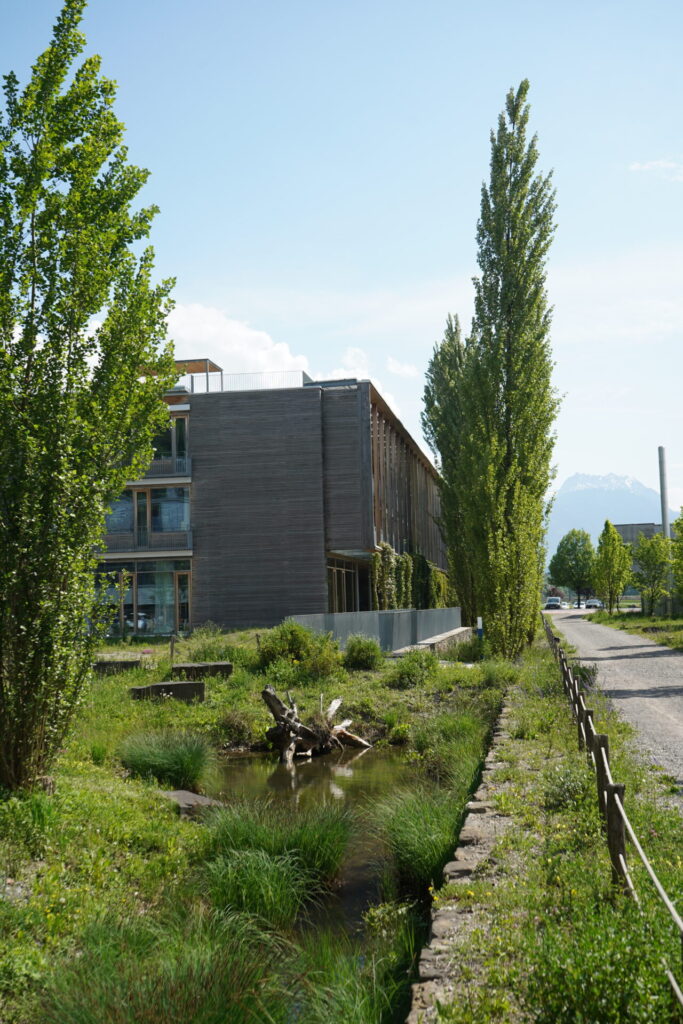 Blick auf ein Firmengelände. Das Unternehmensgebäude ist umgeben von Bäumen, einem Teich und Grünfläche.