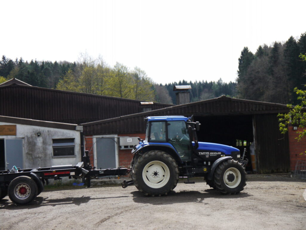 Ein großer Blauer Traktor ist von der Seite zu sehen. Er steht vor einer großen landwirtschaftlichen Halle.
