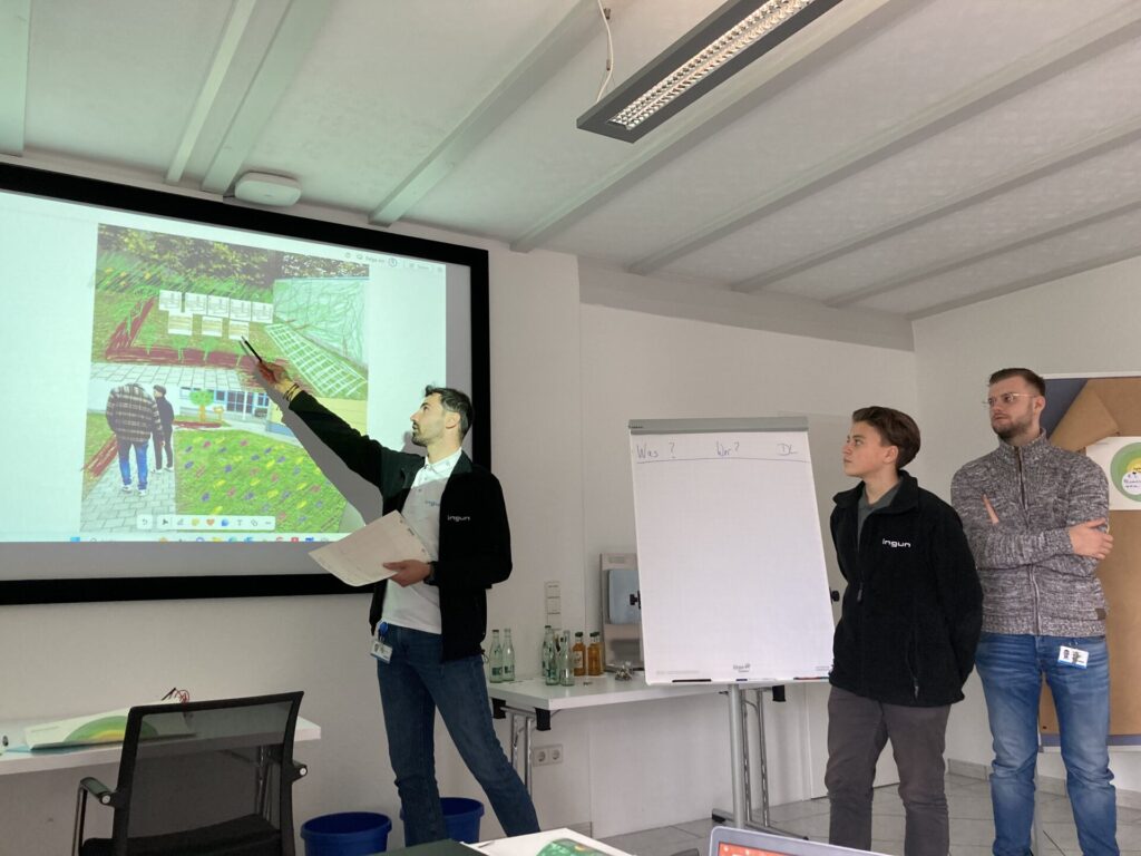 Zwei junge Männer stehen vor einer Leinwand und einem Flipchart. Ein Mann zeigt auf einen gezeichneten Gartenplan auf der Leinwand.