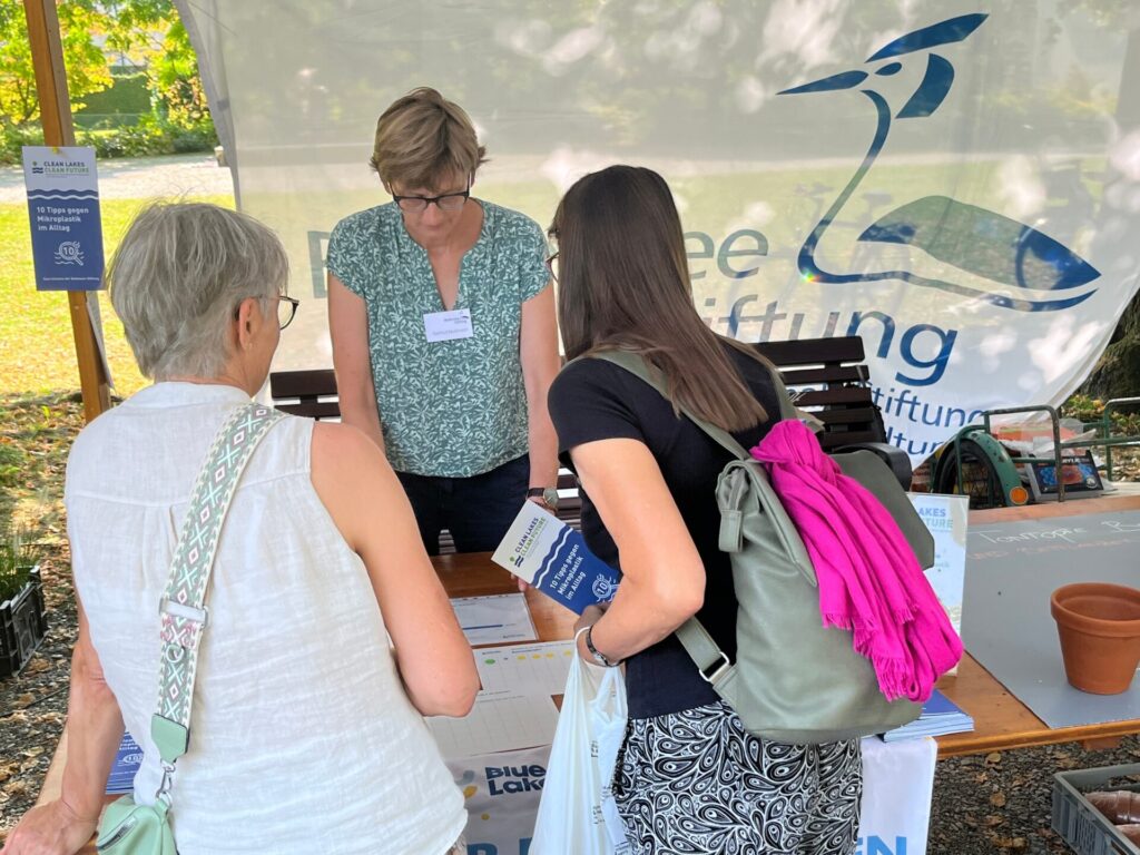 Gertrud Nußbaum steht am Infostand der Bodensee-Stiftung. Vor ihr stehen zwei Frauen mit dem Rücken zur Kamera. Sie halten Info-Flyer in der Hand.