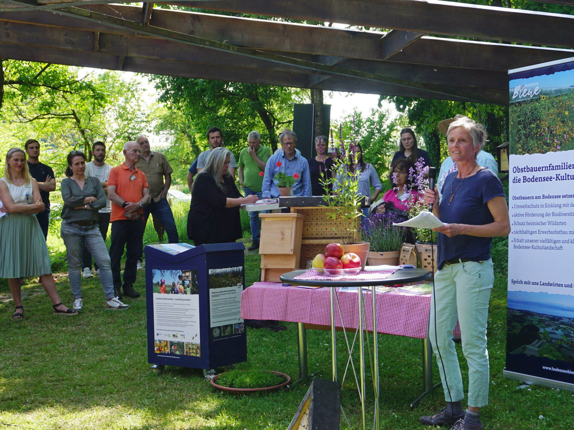Eine Menschengruppe steht im Freien unter einem Dach und blickt auf eine Rednerin. Die Rednerin steht neben einem Tisch mit Obstprodukten.