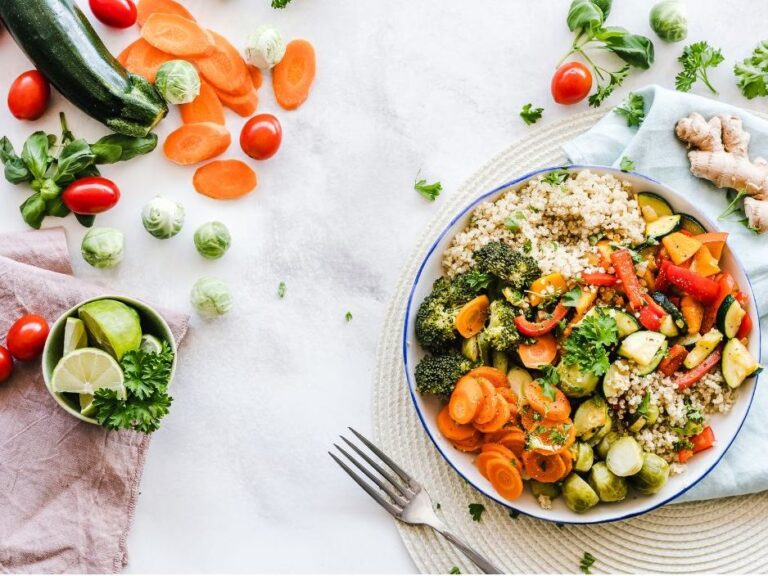 Blick von oben auf einen Tisch mit weißer Tischdecke. Auf einem weißen Platzdeckchen steht ein Teller, der mit buntem Gemüse gefüllt ist. Zucchini, Karotten und Kräuter liegen auch auf der Tischdecke.