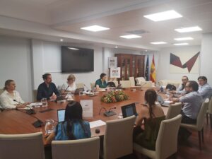 Zusehen ist ein großer runder Tisch, an dem 9 Leute sitzen.